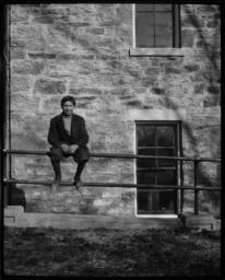 Young Man Sitting on a Fence in front of a Stone Building, Haskell Institute, Lawrence, Kansas
