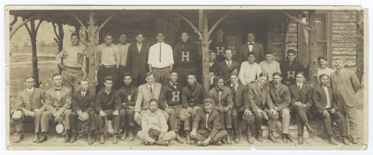 Men in Suits or "H" Sweaters in front of a Log Cabin at the First National YMCA Training Camp