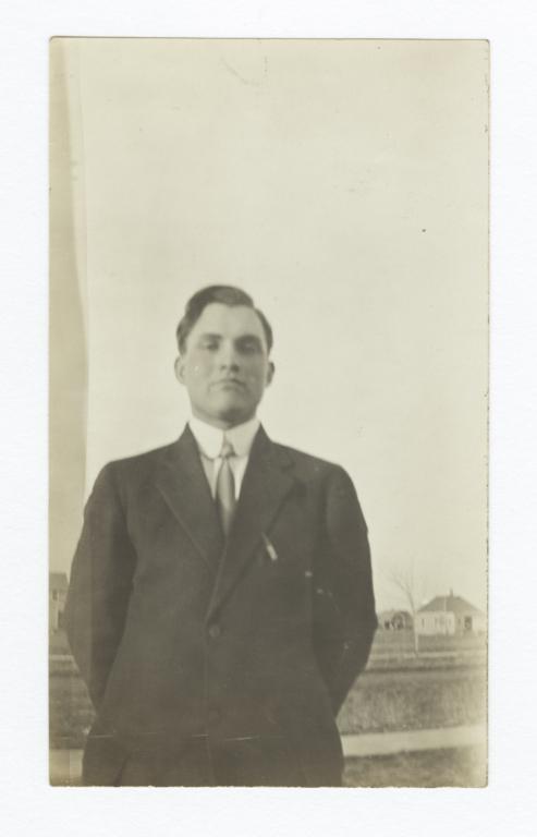 Man Poses Standing Wearing a Suit and Tie on a Field with Small Buildings in the Background  